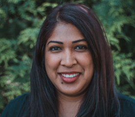A woman with long dark hair and a dark top smiles at the camera. There is a green, leafy background behind her.