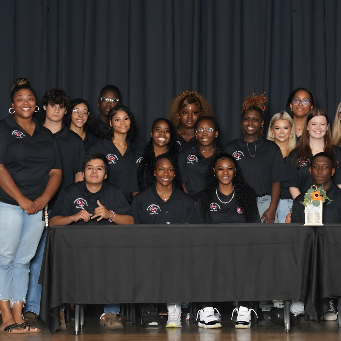 A group of students in matching black shirts pose on stage behind tables with a woman speaking at a podium on the left. A flower vase and sign reading Key to Career are on the table. A dark curtain is in the background.