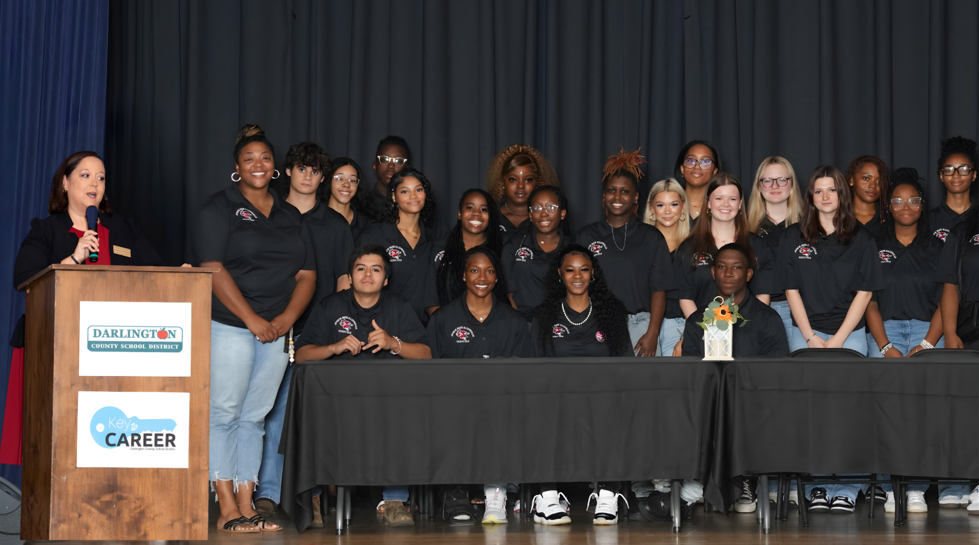 A group of students in matching black shirts pose on stage behind tables with a woman speaking at a podium on the left. A flower vase and sign reading Key to Career are on the table. A dark curtain is in the background.