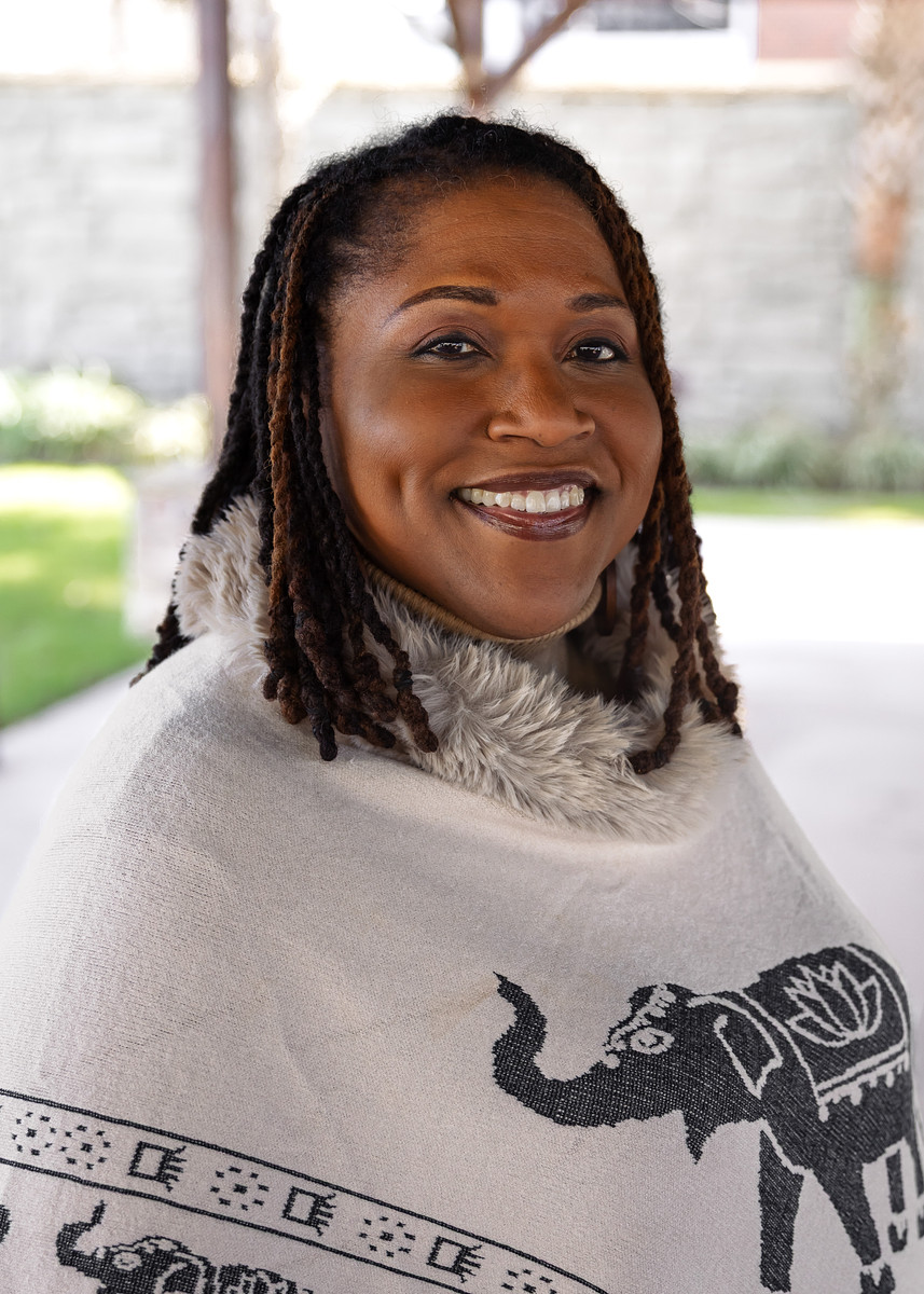 A smiling woman with long, brown, twisted hair wears a white poncho with a faux fur collar and an elephant design, standing outdoors with a blurred background of greenery and a stone wall.
