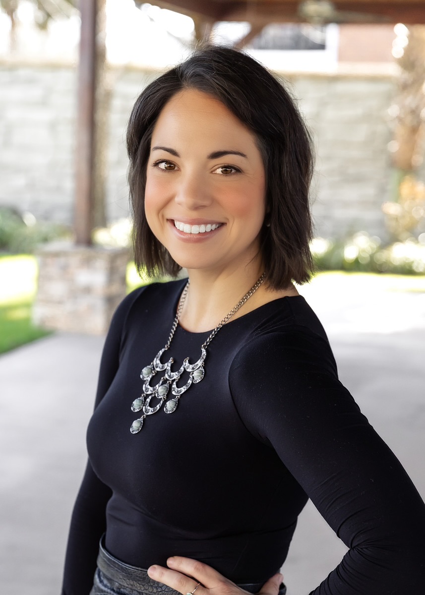 A woman with short dark hair smiles at the camera. She is wearing a black long-sleeve top and a statement necklace. The background is outdoors with blurred greenery and a stone wall.