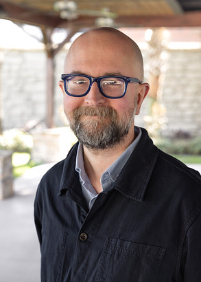 A bearded man with glasses and a shaved head, wearing a dark jacket over a collared shirt, stands outdoors under a covered patio, smiling at the camera. The background is softly blurred.