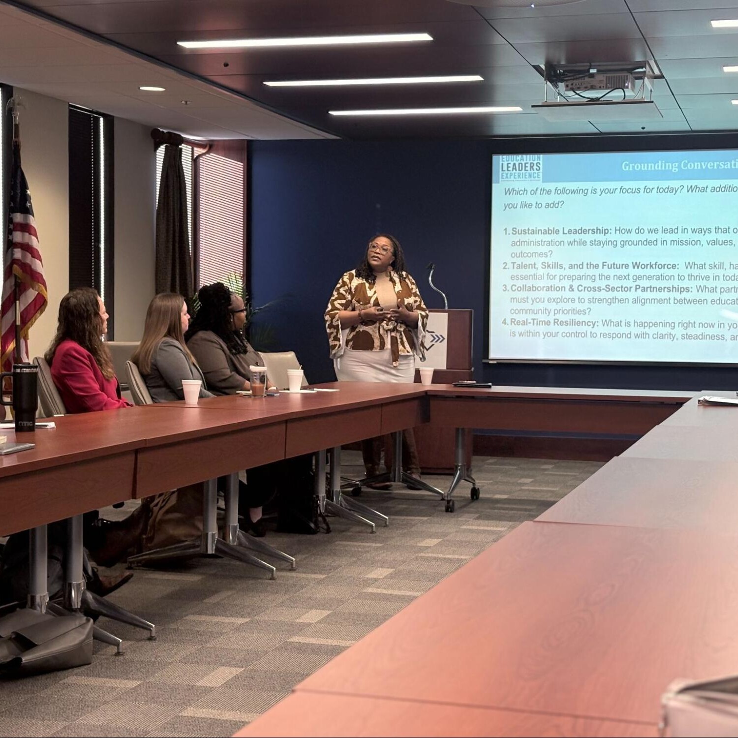 A woman stands and presents to a group seated around a conference table in a meeting room. A large screen displays discussion topics such as leadership, talent, collaboration, and resilience. An American flag is visible.