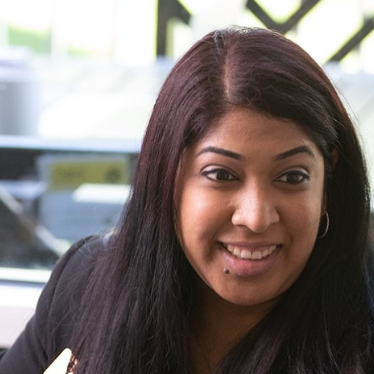 A woman with long dark hair smiles while talking to another person across a desk, with office supplies nearby and a window in the background.