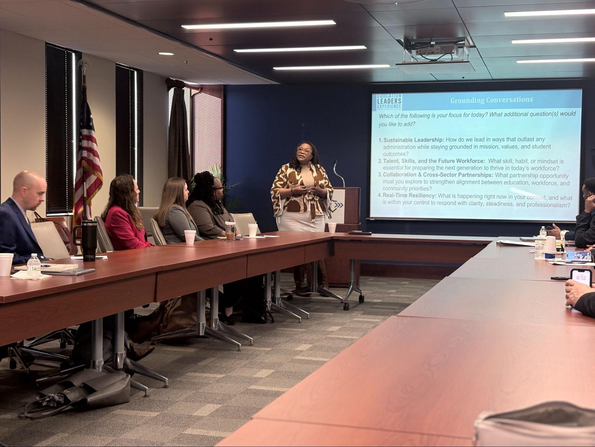 A woman stands and presents to a group seated around a conference table in a meeting room. A large screen displays discussion topics such as leadership, talent, collaboration, and resilience. An American flag is visible.