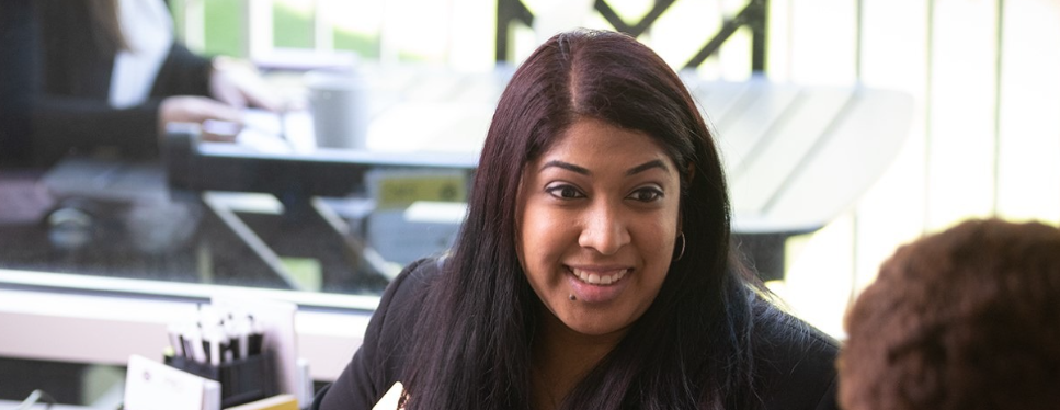 A woman with long dark hair smiles while talking to another person across a desk, with office supplies nearby and a window in the background.