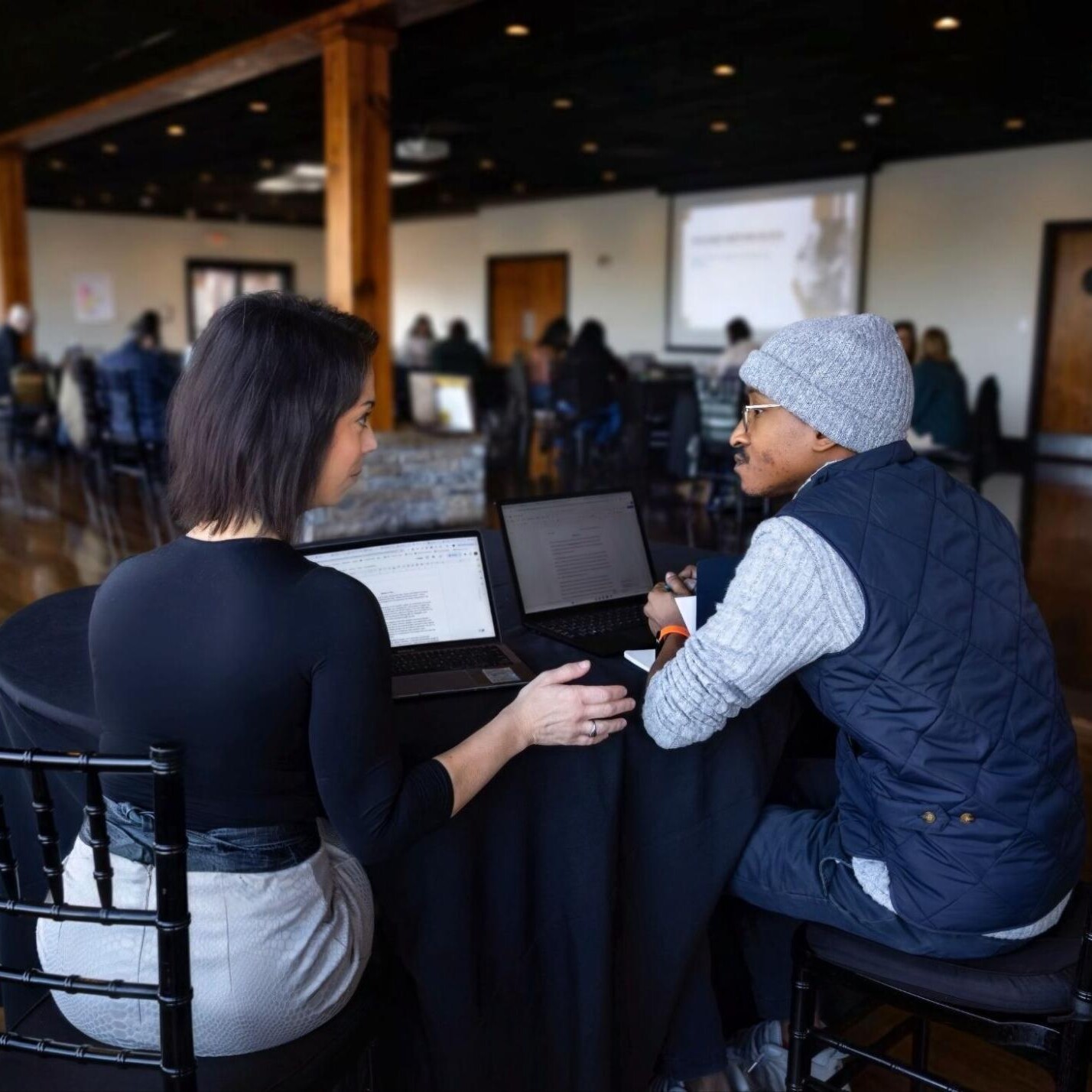 Two people sit at a round table, each with a laptop, engaged in conversation in a spacious, modern room with other people seated in the background, possibly attending a workshop or conference.