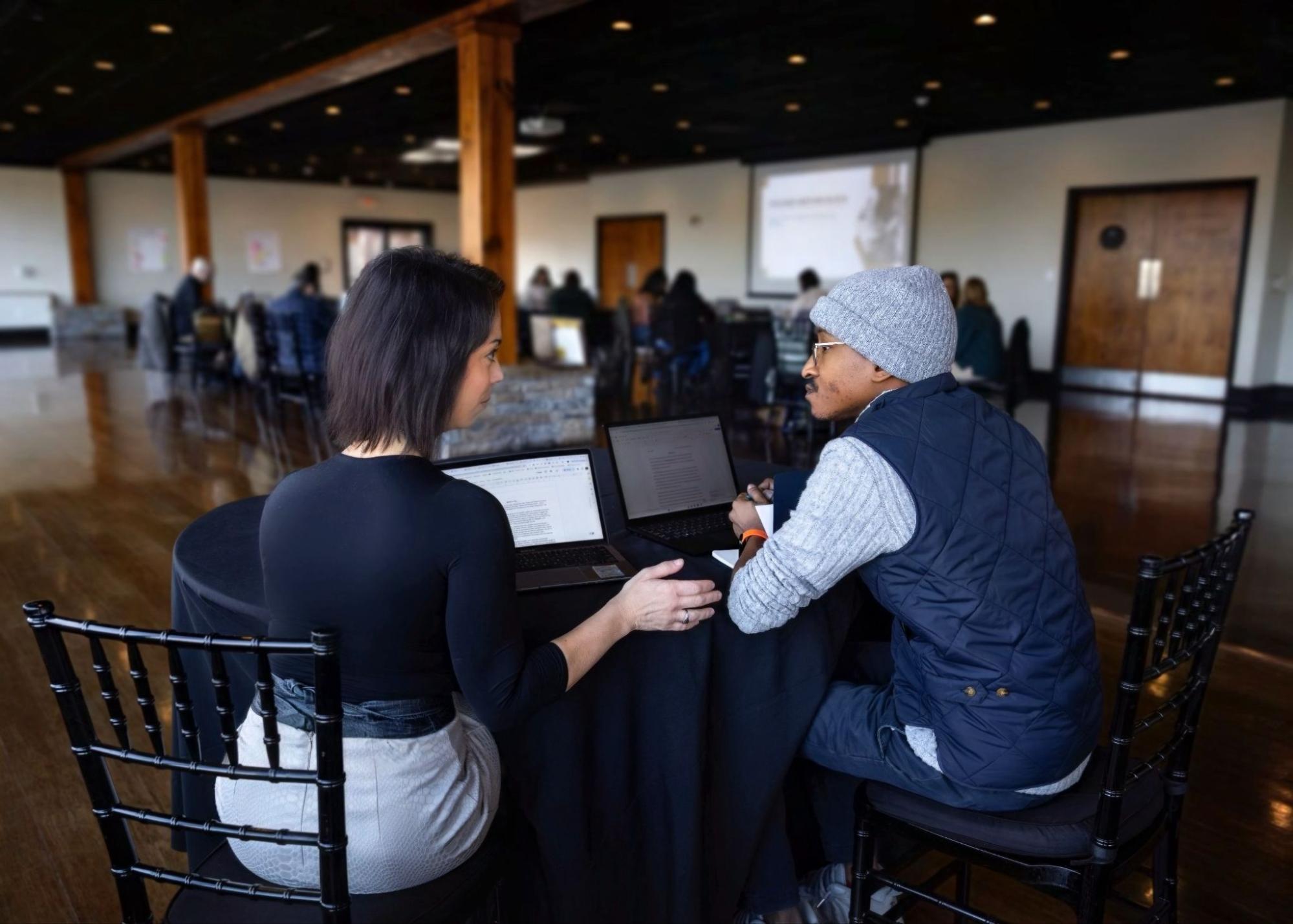 Two people sit at a round table, each with a laptop, engaged in conversation in a spacious, modern room with other people seated in the background, possibly attending a workshop or conference.