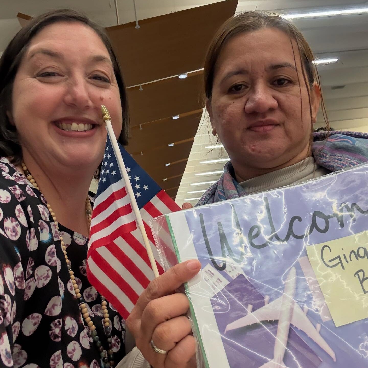 Two women pose for a selfie indoors; one is smiling and holding a small American flag, while the other holds a folder with paper airplanes and a visible Welcome sign.