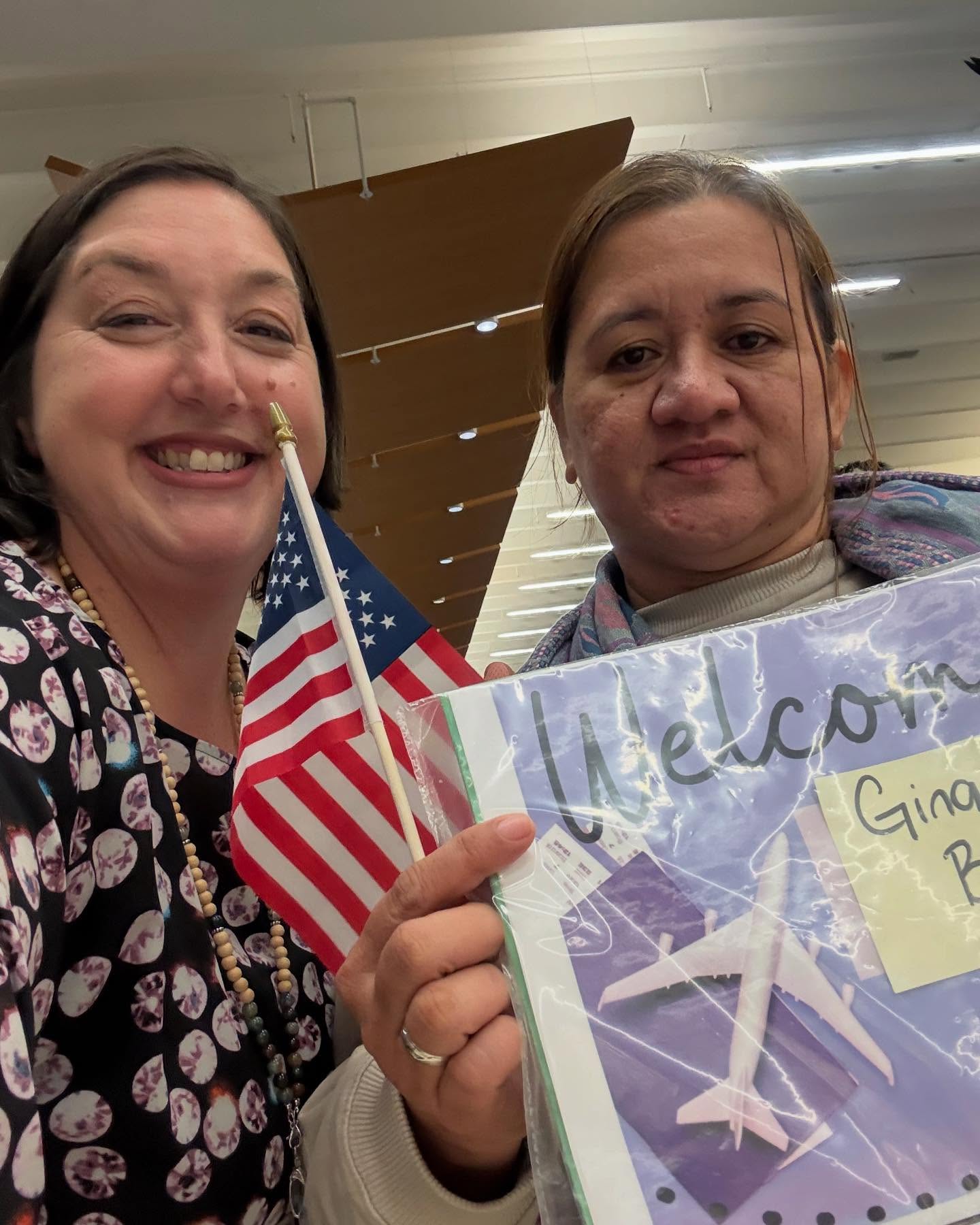 Two women pose for a selfie indoors; one is smiling and holding a small American flag, while the other holds a folder with paper airplanes and a visible Welcome sign.