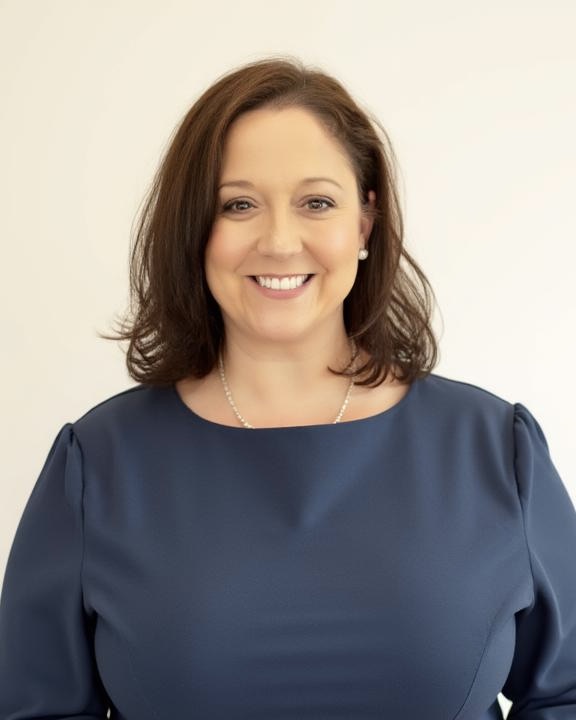 A woman with shoulder-length brown hair, wearing a navy blue dress and a pearl necklace, smiles at the camera against a plain light background.