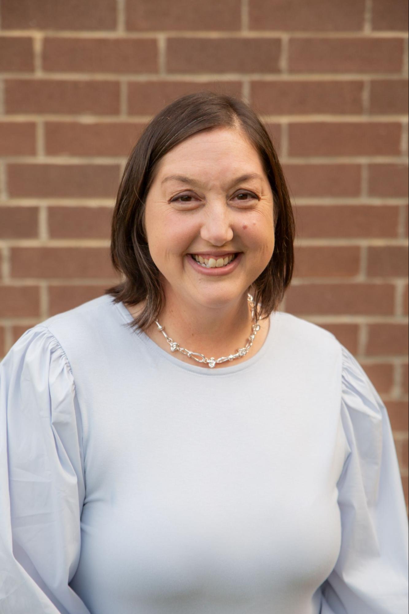 A woman with short brown hair and a light blue blouse smiles at the camera, standing in front of a red brick wall. She is wearing a silver necklace.